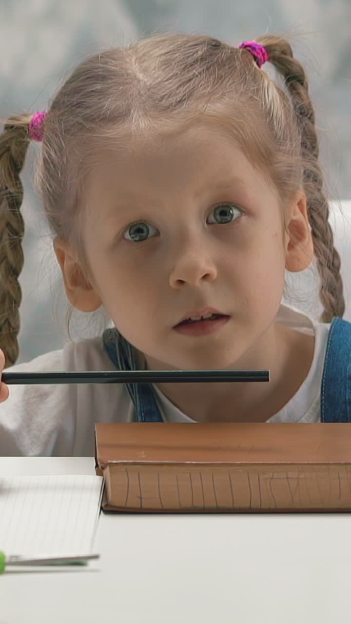 close up slow motion portrait of little girl student with pigtails in sundress, sitting at table at home, whiting, knocking pencil on book, nervous, kid looking for object with her eyes