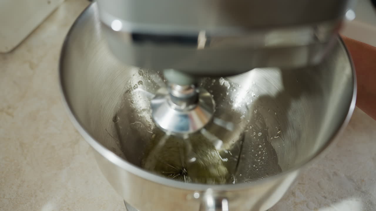 Close up of electric mixer whisking egg fluid in stainless steel bowl on kitchen counter during cooking process showing high-speed rotation and fluid motion inside mixing bowl