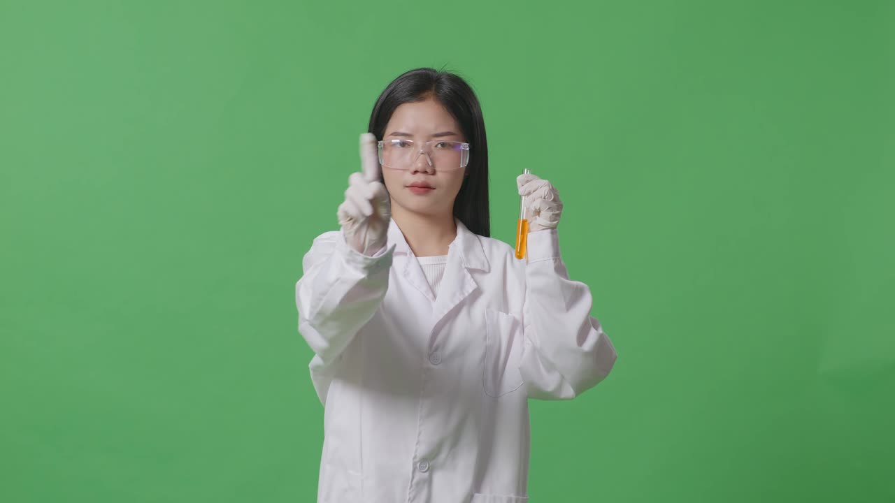 시험관에 오렌지색 액체를 넣은 아시아 여성 과학자가 당신을 가리키고 실험실의 녹색 스크린 배경에 지표 손가락 표시 없이 불쾌감을 표합니다.
