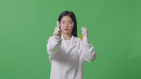 asian woman scientist with orange liquid in the test tube pointing at you and disapproving with no index finger sign on the green screen background in the laboratory