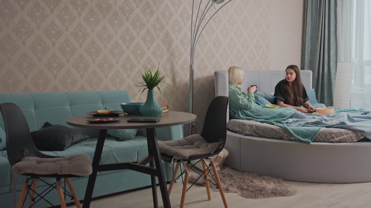 Two young women relax on bed covered with teal blankets, sipping coffee and having friendly conversation in bright cozy bedroom with soft lighting, modern furniture, and casual breakfast setup