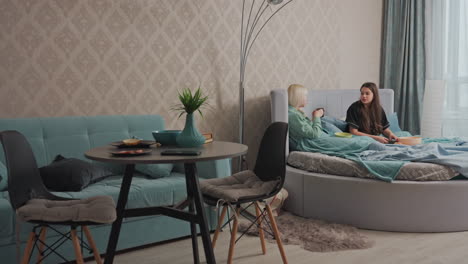 two young women relax on bed covered with teal blankets, sipping coffee and having friendly conversation in bright cozy bedroom with soft lighting, modern furniture, and casual breakfast setup