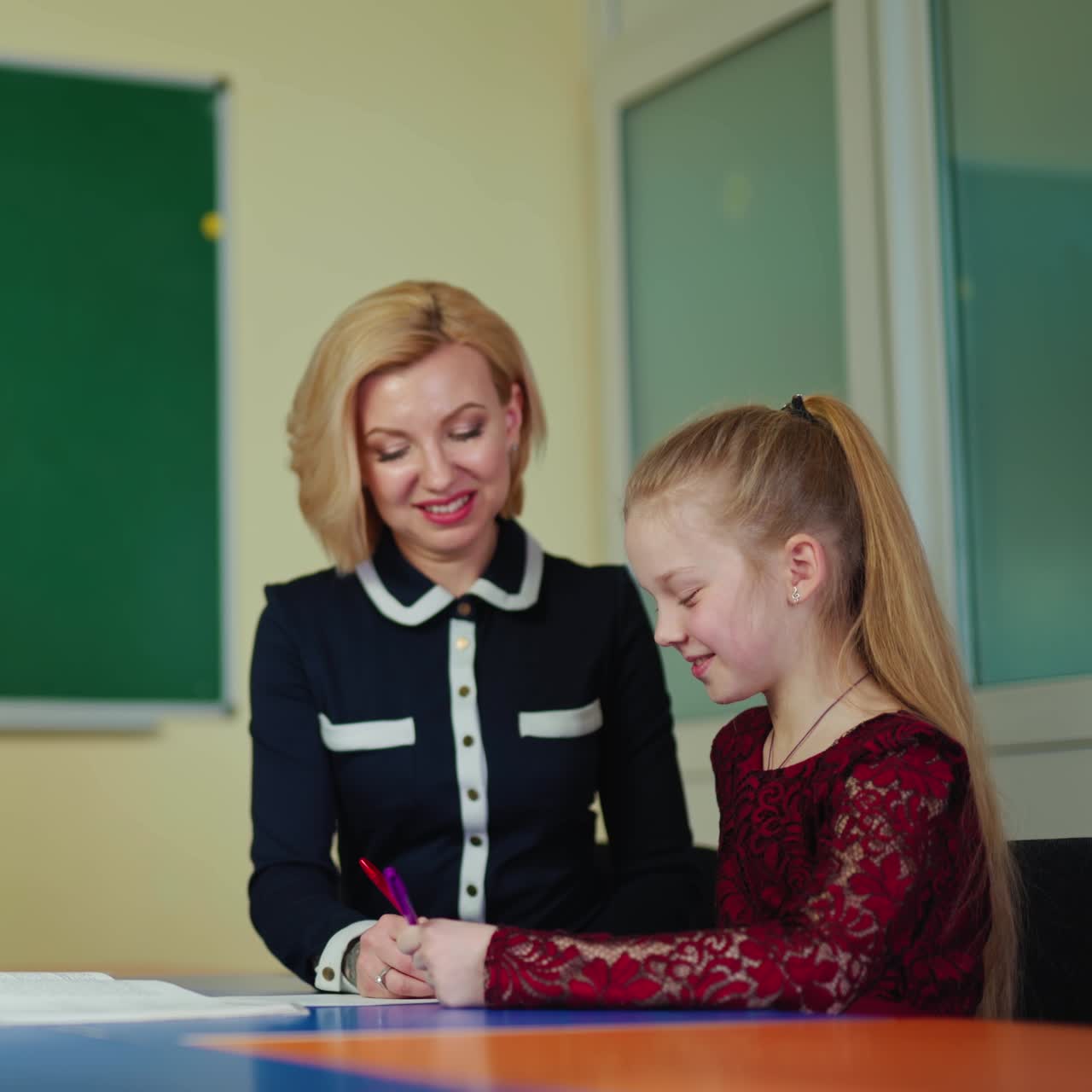 Positive teacher with a cute schoolgirl. Little student learning writing with a tutor. Experienced teacher helps studying elementary pupil at school table.