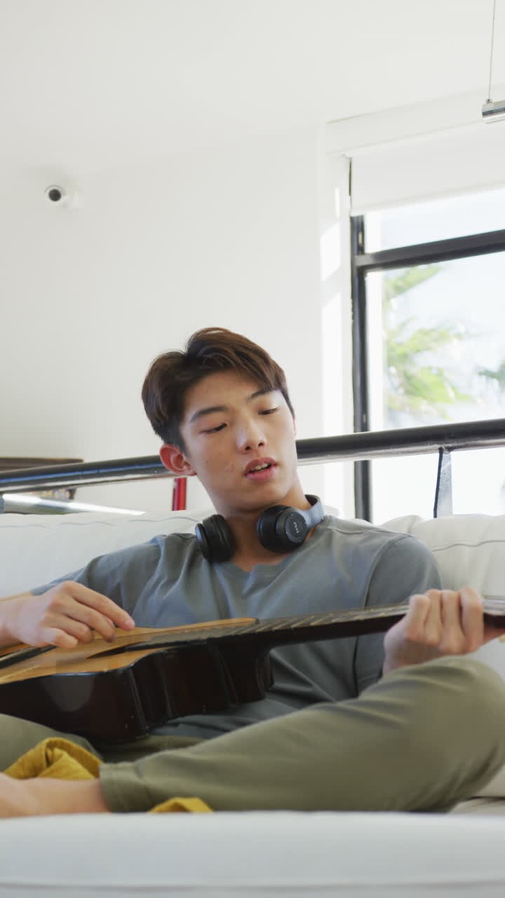 video verticale di un adolescente asiatico che suona la chitarra in soggiorno