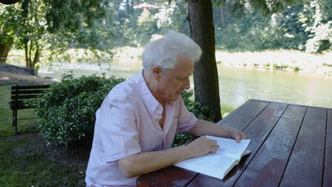 older man reads book at table by river, laughing, enjoying, trees