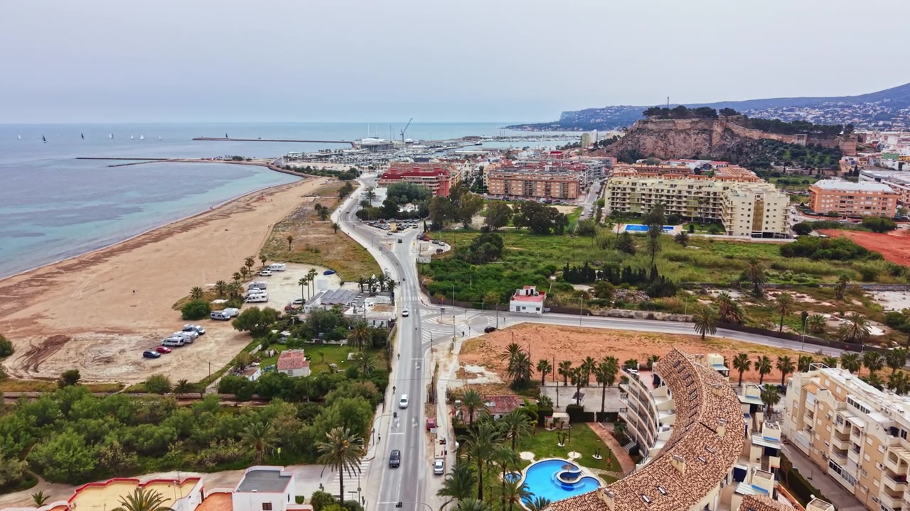 Kystområde viser bygninger, veje og strand med hav i baggrunden på en overskyet dag.