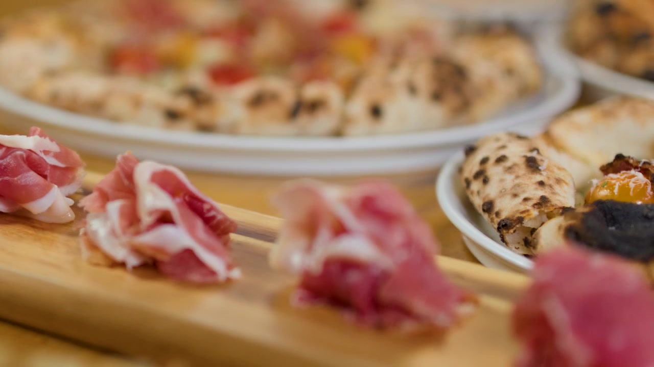 Primer plano en movimiento de aperitivos de embutidos con pizza desenfocada al fondo.