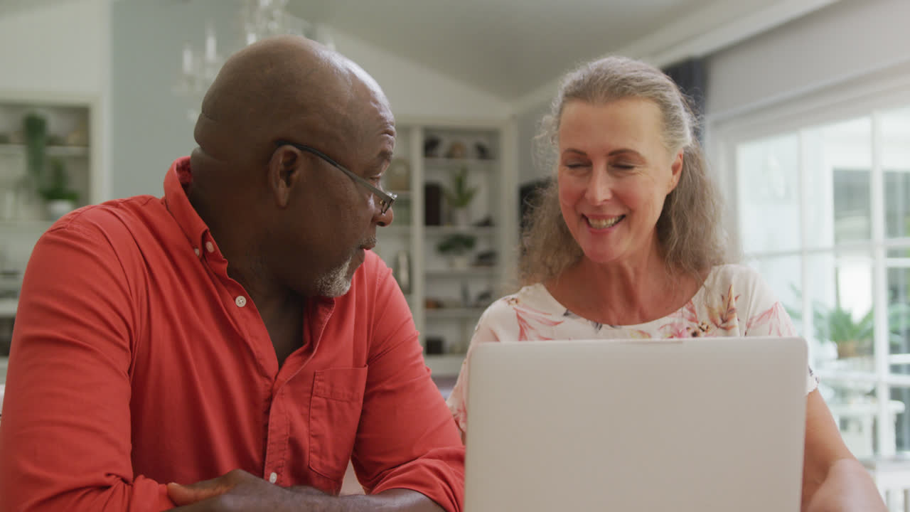 Happy senior diverse couple wearing shirts and using laptop in living room