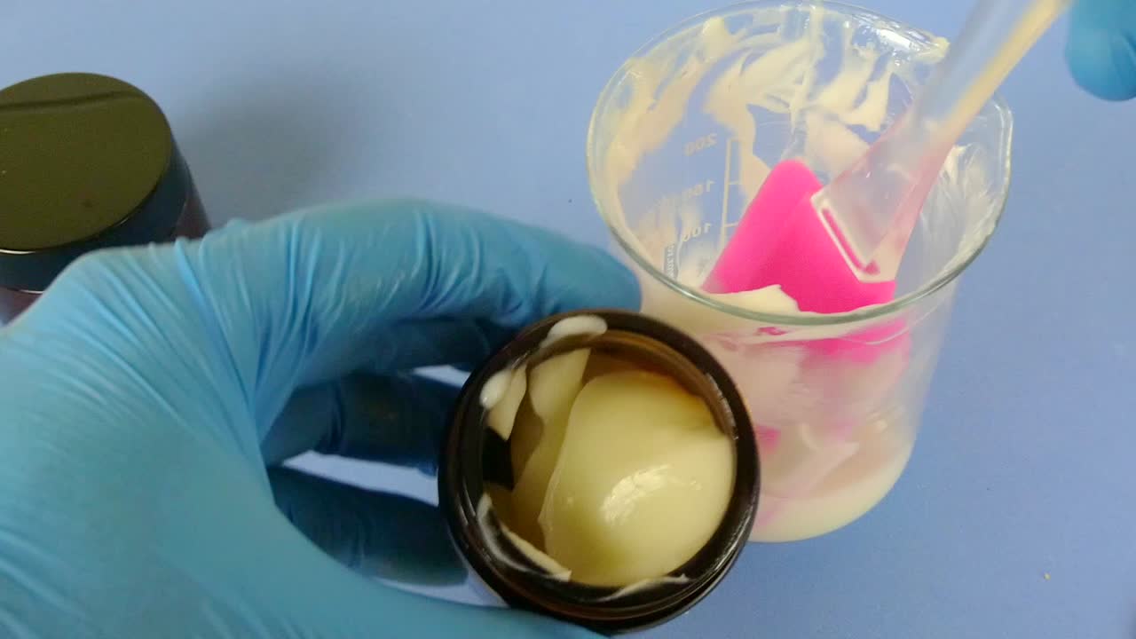 Macro view of a cosmetic laboratory scene where a gloved operator uses a spatula to carefully package a white emulsion.