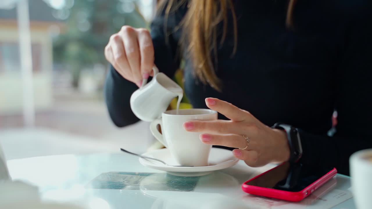 Woman pouring milk into coffee cup on the table. Hands of a young lady adding hot milk to coffee in a cafe. Close-up. Slow motion.