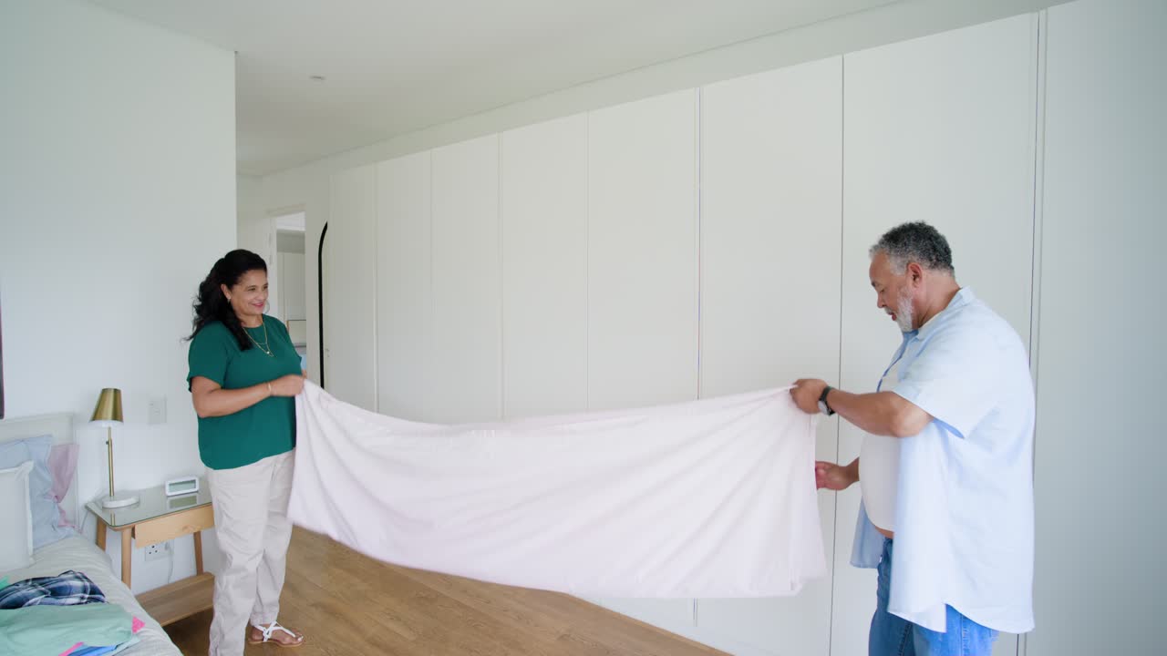 Coppia afroamericana, la donna prende un lenzuolo rosa pallido, piegandolo per togliere le pieghe in camera da letto.