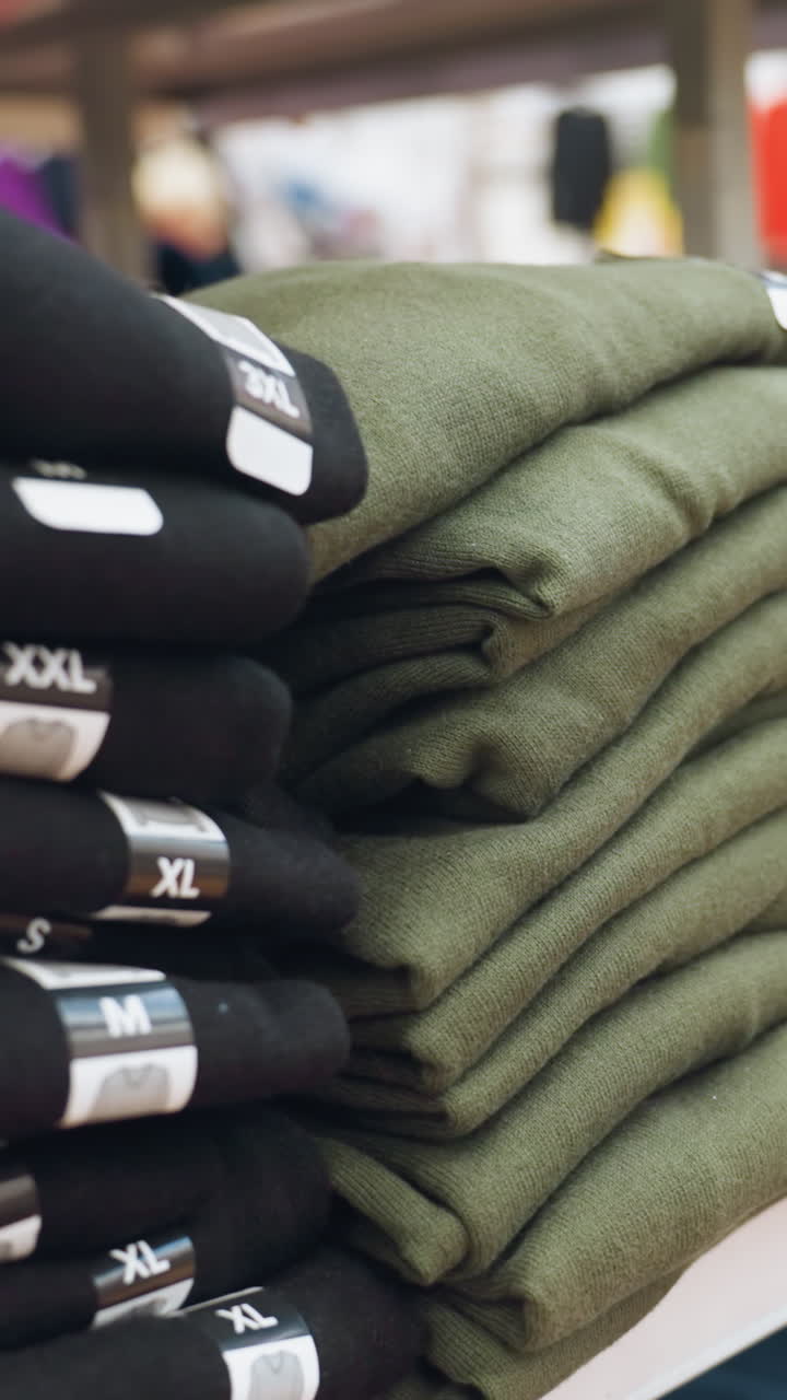 view of neatly folded clothes in various colors with size tags (S, M, L, XL, XXL) displayed on shelves in a mall. Captured with a handheld camera, emphasizing the organized retail environment