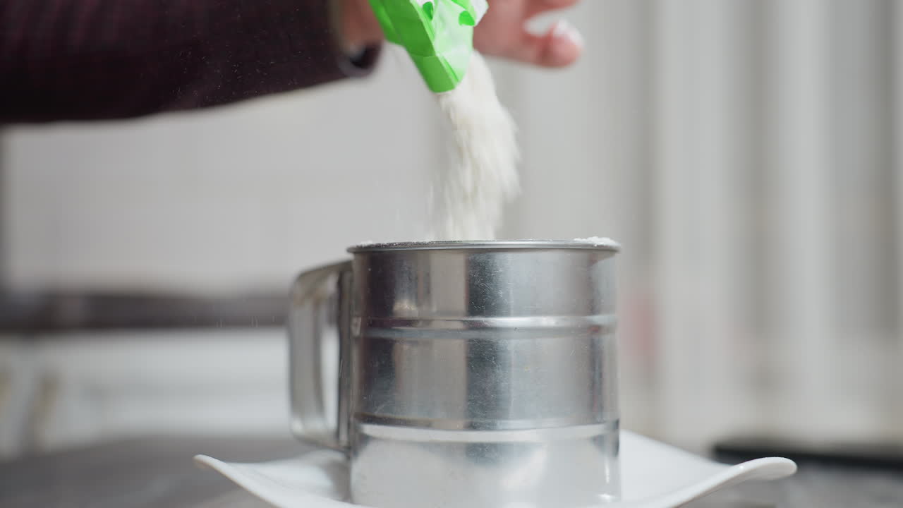 Close-up of person pouring flour into a metal sifter cup placed on a white plate, the motion of the fine powder creates a soft flow, emphasizing the precision of baking preparation
