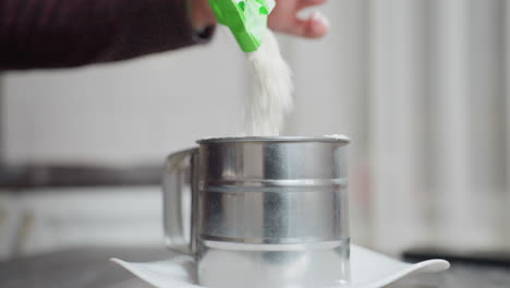 primer plano de una persona vertiendo harina en una taza de tamiz de metal colocada en una placa blanca, el movimiento del polvo fino crea un flujo suave, haciendo hincapié en la precisión de la preparación del pan