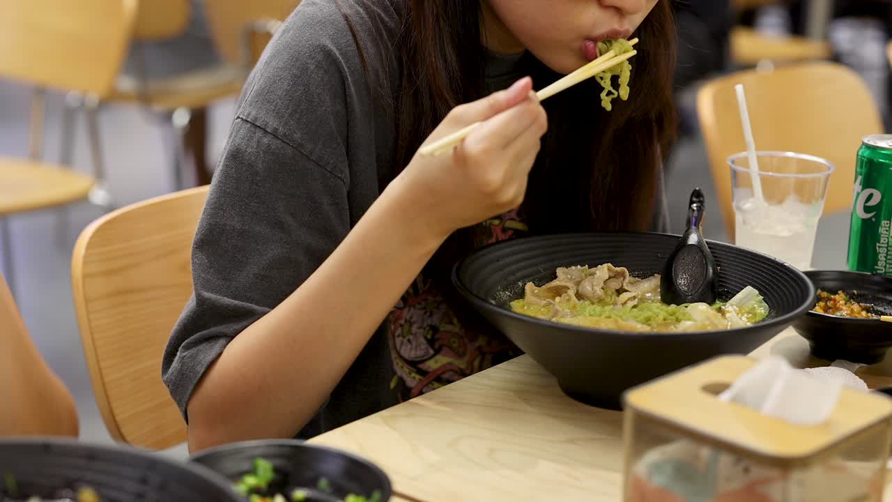 Young woman eats green noodles with chopsticks in brightly lit, casual Bangkok food court setting
