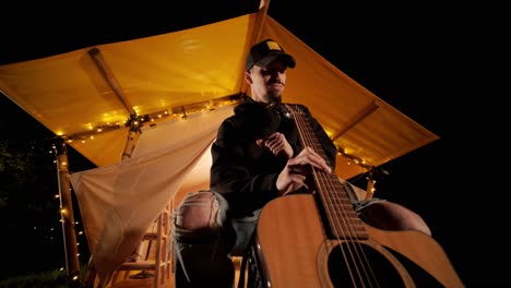 man plays the guitar and sings songs in a tent with light bulbs