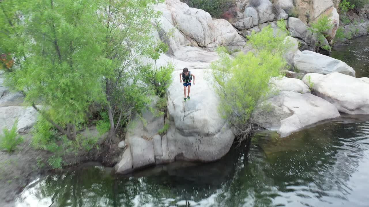 A drone flies backward as a young boy enjoys jumping off a cliff.