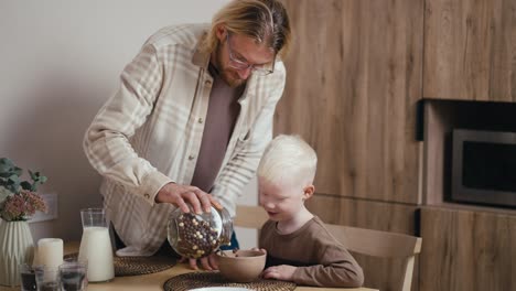 ein glücklicher blonder mann mit brille und bärte gießt seinem kleinen albinen sohn mit weißen haarfarbe in einer schüssel verschiedene farbige müslchen und fügt dann milch hinzu und macht morgens frühstück für seinen kleinen sohn in einer modernen küche