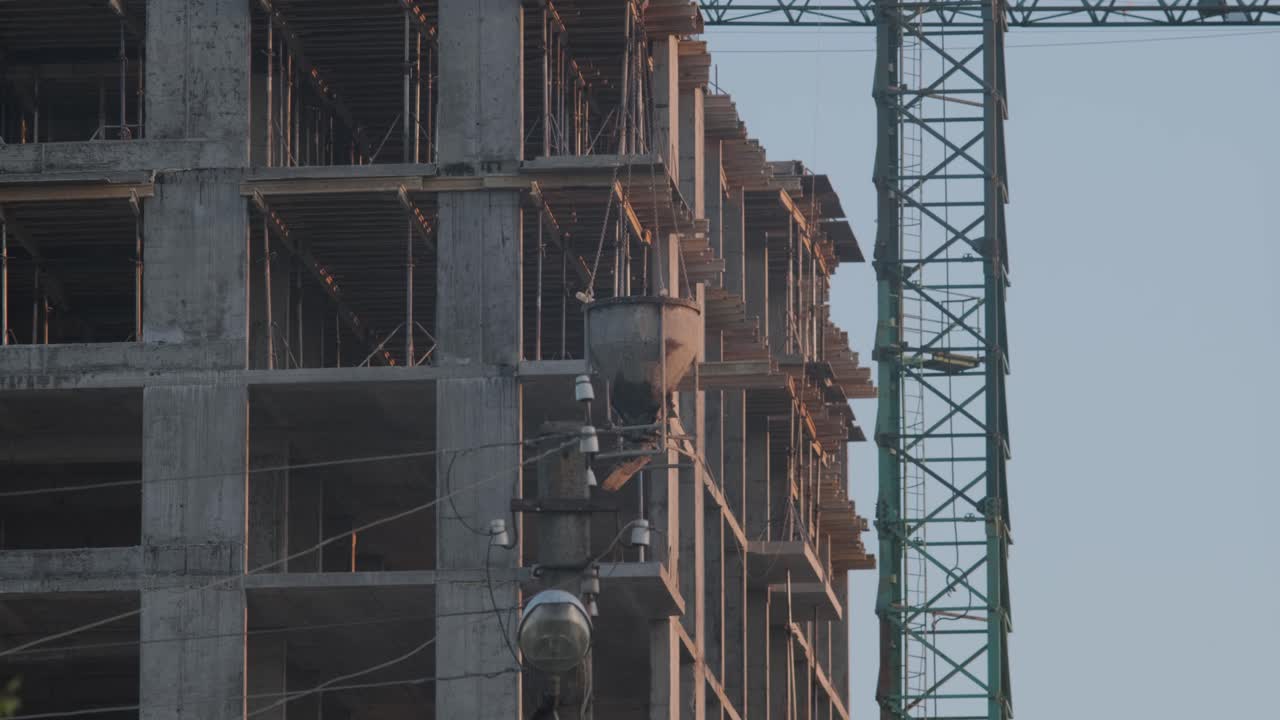 un primer plano de un edificio de gran altura en construcción, con estructuras de hormigón visibles y andamios, al lado de una grúa, capturado al anochecer