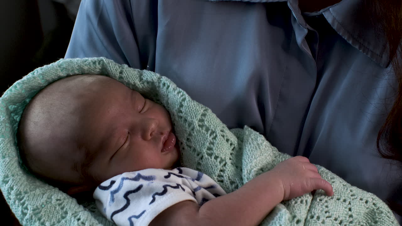 Un primo piano di un bellissimo neonato cullato tra le braccia della madre, avvolto in una coperta, con gli occhi chiusi mentre dorme serenamente
