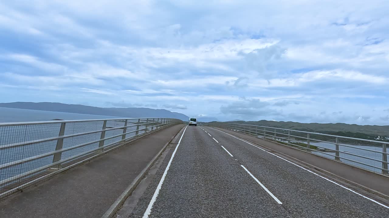 Forward-facing dashcam view of a vehicle crossing a long bridge over water toward the Isle of Skye, with overcast skies and steady camera movement