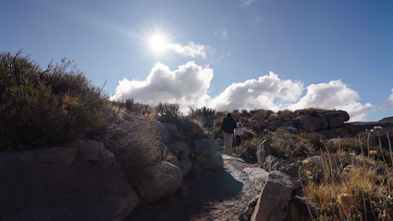 Still shot of a mother and daughter hiking and walking uphill trail surrounded by bushes with bright sun, Malargue, Mendoza, Argentina