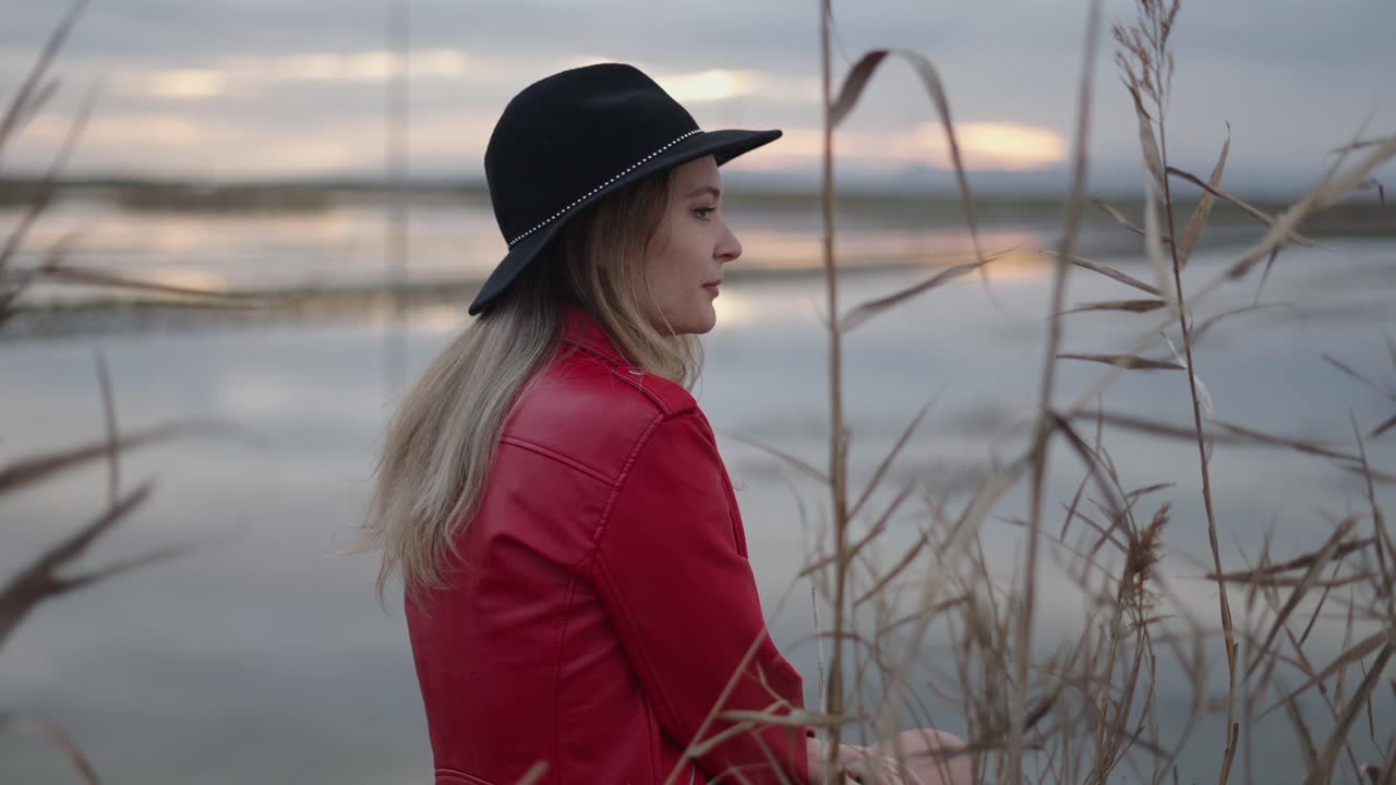Woman in Red Jacket and Black Hat by the Water at Sunset