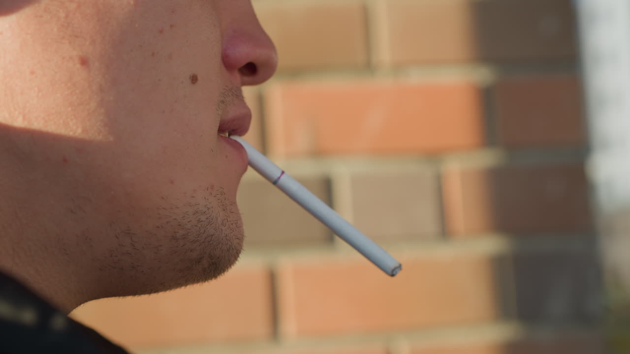 close up side view of man holding cigarette between lips preparing to light it outside with blurred brick wall background under sunlight during daytime