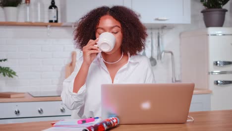 joven mujer afroamericana bebe café se sienta en la mesa con una computadora portátil en la cocina