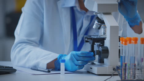 primer plano de un trabajador de laboratorio mirando a través del microscopio y registrando los resultados de las pruebas en la computadora