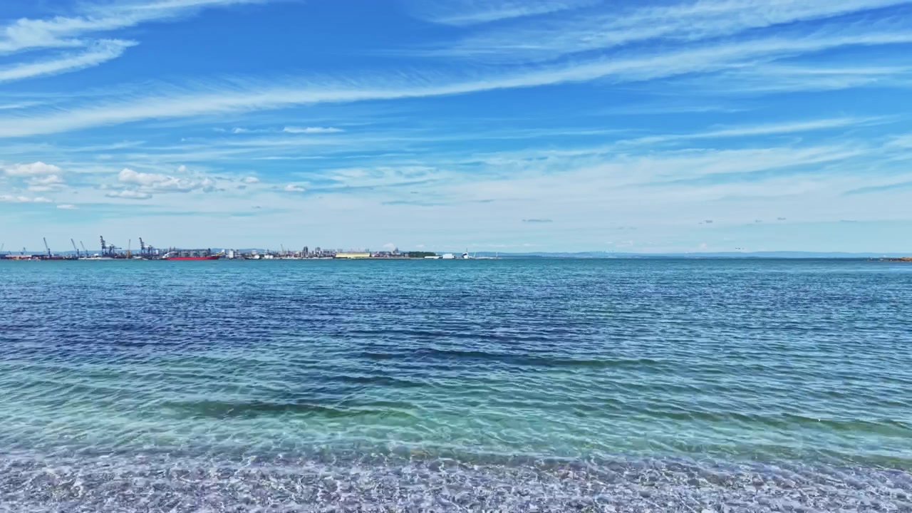 Friedlicher Blick auf Meer und Himmel. Ruhiges Wasser verschmilzt mit der fernen Stadtsilhouette unter sanftem Himmel. Sanfter Meereshorizont mit stillen Gewässern und schwebenden Wolken über der Stadtlandschaft.