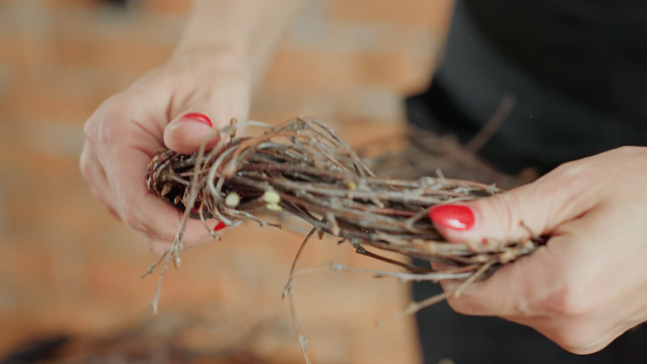 Female hands holding and arranging dry twigs while crafting rustic wreath on straw base, focusing on handmade decoration, detailed artisan process, and natural materials for seasonal home decor