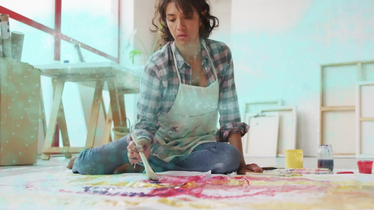 Woman artist sitting on studio floor, camera focusing on brush, dipping paint, refining artwork