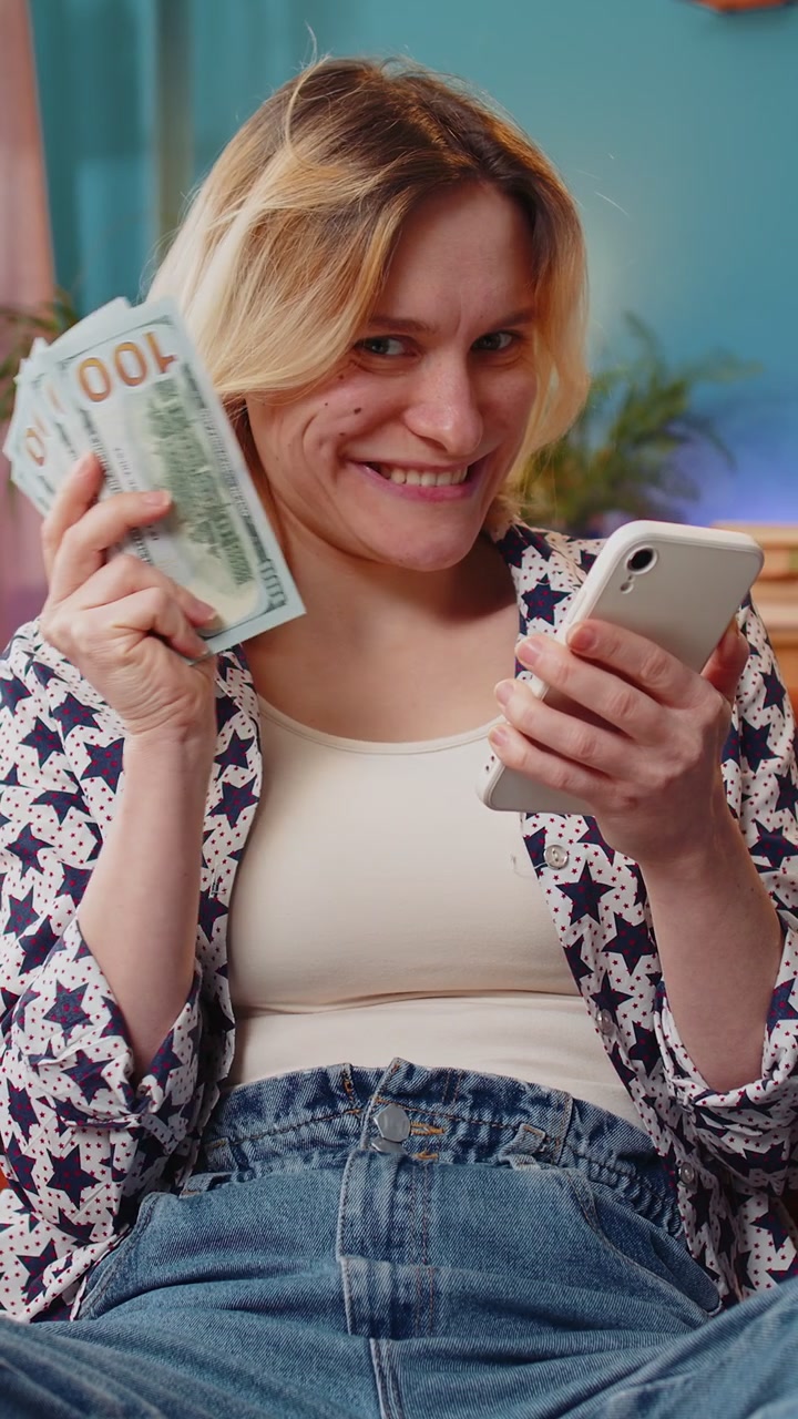 Succesvolle rijke glimlachende vrouw met een smartphone die dollarbiljetten zwaait, een geldfan die op de thuissofa zit.
