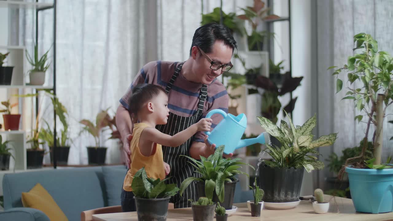 아기 와 함께 아시아인 남자 가 집 에 있는 식물 들 에게 물 을 주기 위해 물병 을 들고 있다