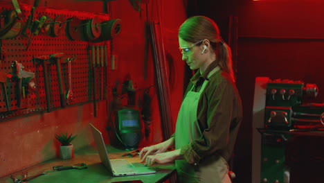 Side-view-of-young-woman-in-apron-and-goggles-using-laptop-in-metal-workshop