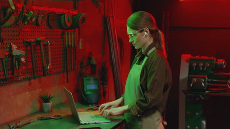 vista lateral de una joven con delantal y gafas usando una laptop en un taller metalúrgico