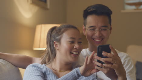Smiling-Asian-Couple-Relaxing-On-Sofa-At-Home-Looking-At-Mobile-Phone-In-Evening-Together