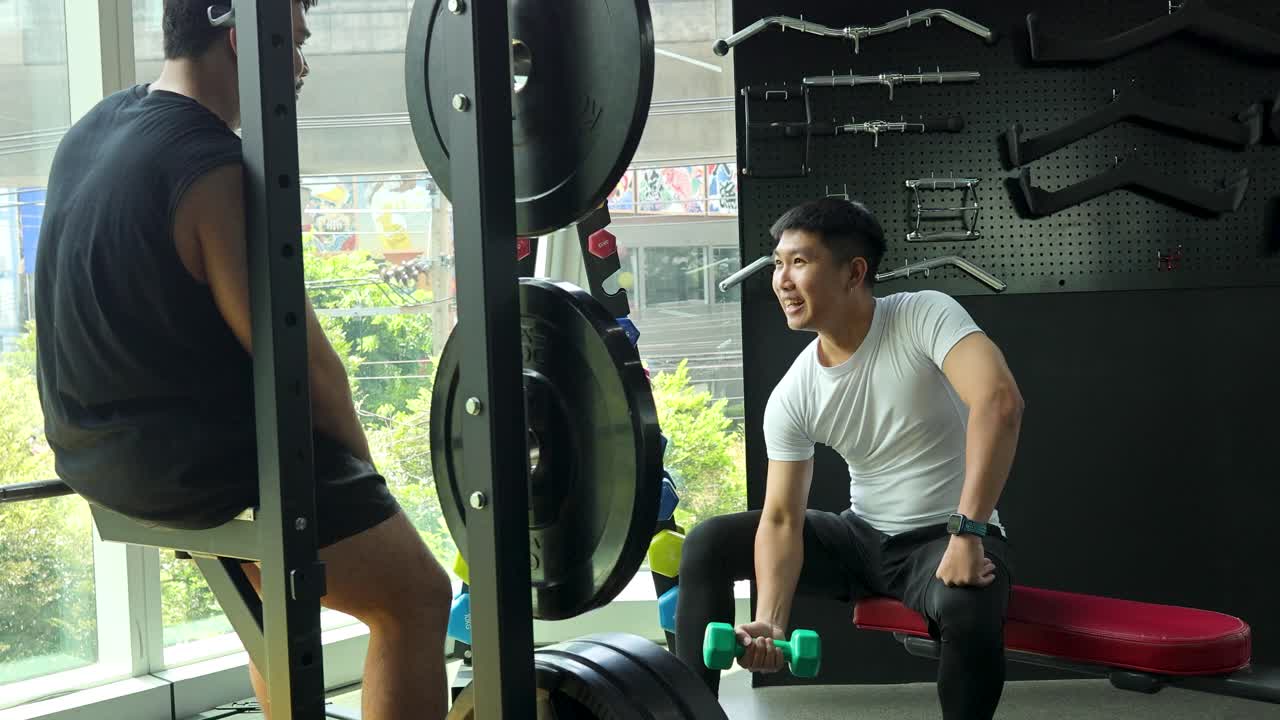 Two men in a modern gym, one seated lifting dumbbells, conversing in bright natural daylight
