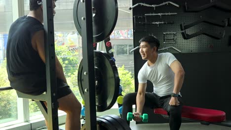 two men in a modern gym, one seated lifting dumbbells, conversing in bright natural daylight