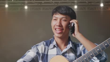 primer plano de un adolescente asiático con una guitarra hablando en su teléfono inteligente mientras está de pie en el escenario