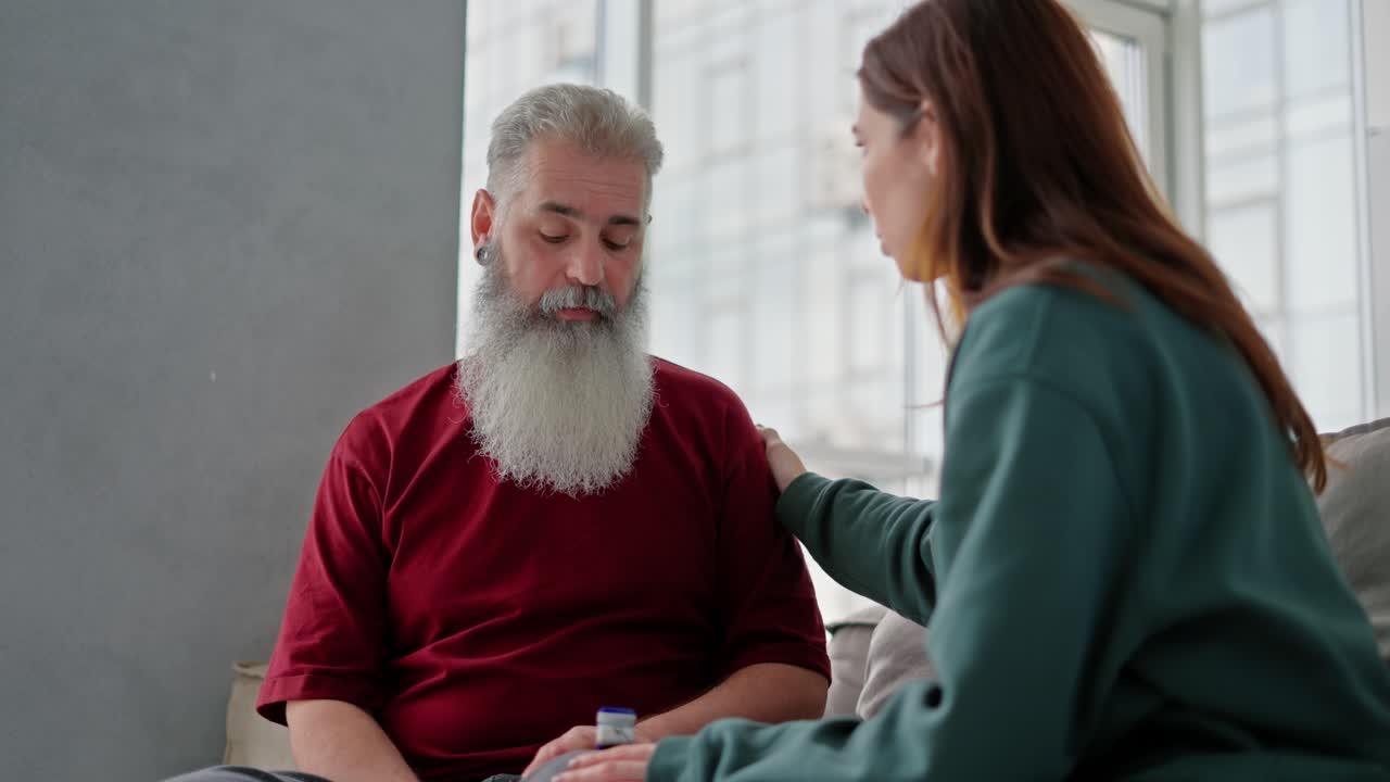 An adult brunette girl in a green jacket talks with her dad an elderly man with gray hair and a lush beard in a red T-shirt bout his health in a modern apartment