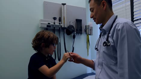 Side-view-of-young-asian-male-doctor-showing-syringe-to-caucasian-boy-patient-in-clinic-4k