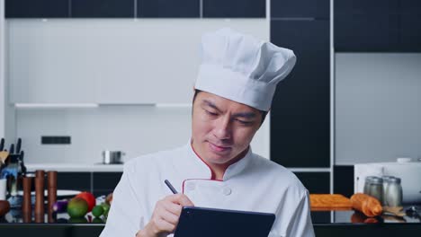 close up de un chef asiático tomando nota en una tableta mientras está de pie en la cocina de casa