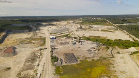 aerial drone footage flying around a large limestone quarry mine where conveyor belts, machines, equipment and mining roads are visible on the dusty ground between waste water puddles at sunny day