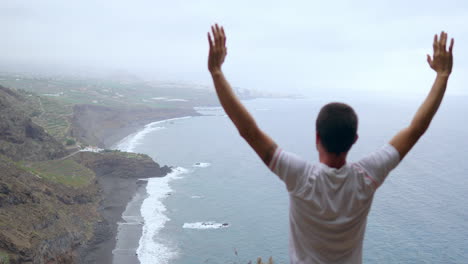 Mit-Dem-Meer-Vor-Sich-Hebt-Ein-Mann-Am-Rand-Einer-Klippe-Die-Arme,-Atmet-Während-Einer-Yoga-Übung-Die-Meeresluft-Ein-Und-Findet-Gelassenheit