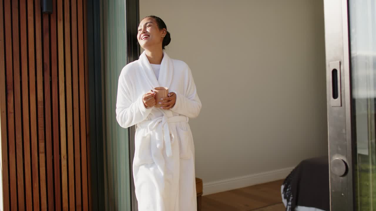 Une femme asiatique en robe, guidée par la lumière du soleil chez elle, lève une tasse et sourit face à la porte coulissante tout en admirant la vue