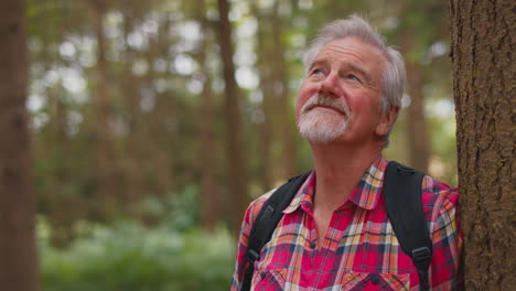 active retired senior man walking in woodland countryside wearing backpack leaning  on tree