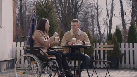 niña discapacitada en silla de ruedas y su amigo bebiendo batido y hablando juntos mientras están sentados en la terraza de un bar