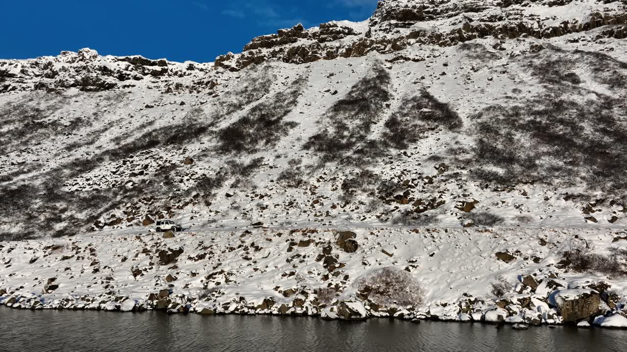 İzlanda'daki Kleifarvatn'ın karlı uçurum kenarındaki kıyısı boyunca ilerleyen beyaz bir 4x4'ün havadan yan görünümü, aşağıda kayalık yamaçlar ve karanlık kış suyu ile.